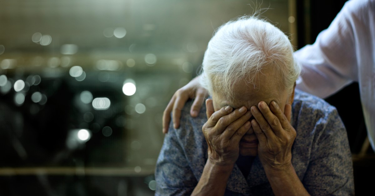 Een oudere vrouw heeft een gezondheidsprobleem, haar hoofd wordt ondersteund door haar armen en iemand houdt haar schouders vast.