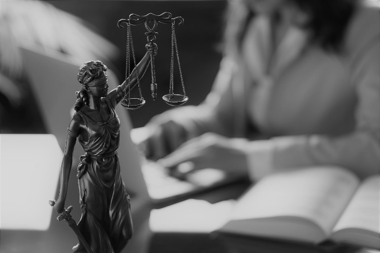 een vrouw zittend aan een tafel met een boek voor zich en een standbeeld van rechtvaardigheid op de voorgrond op de tafel