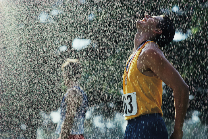 Een mannelijke atleet na het passeren van de finish. Het regent. Zijn hoofd is omhoog gedraaid tegen de vallende druppels. Zijn ogen zijn gesloten, hij draagt een medaille om zijn nek. Hij draagt een gele tanktop met het nummer 63 erop en een blauwe korte broek.