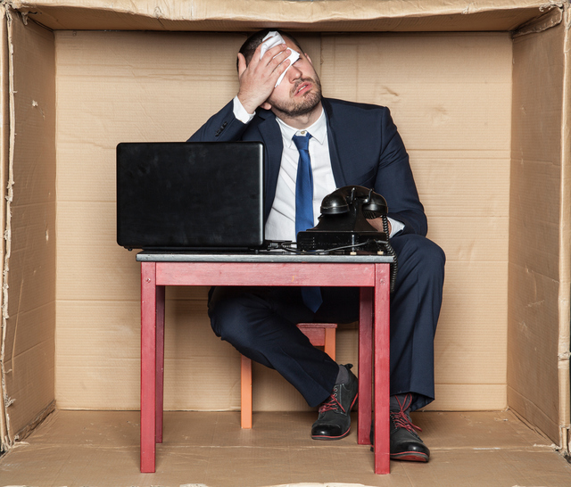 Zakenman zittend aan een bureau in een doos, krappe ruimte, werkstress en verhoogde werkdruk, computer en telefoon, man veegt zijn voorhoofd af