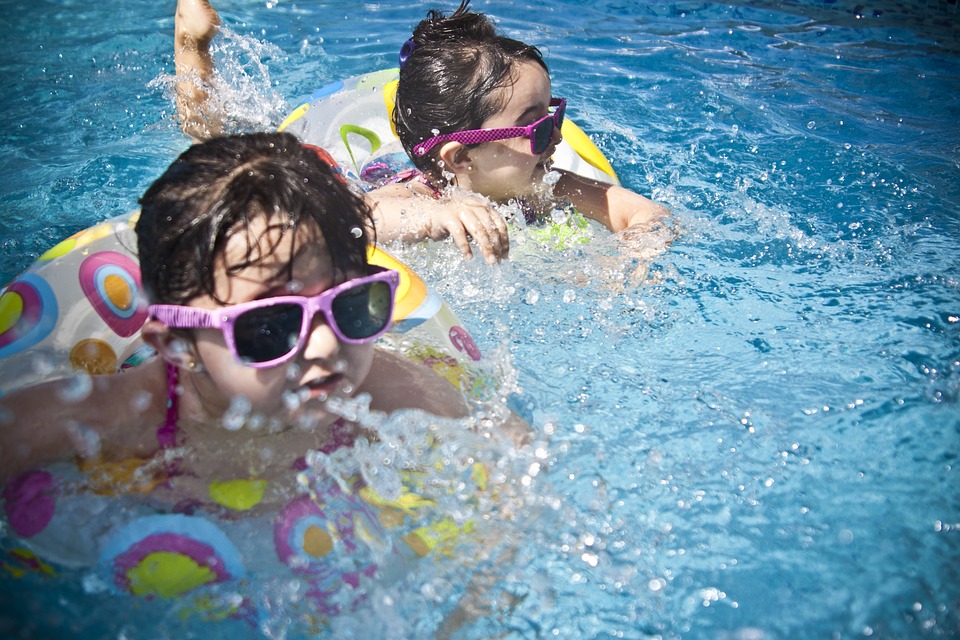 Twee kinderen zwemmen in het water. Ze dragen een zonnebril met een roze montuur. Ze zitten in drijvende fietsen.