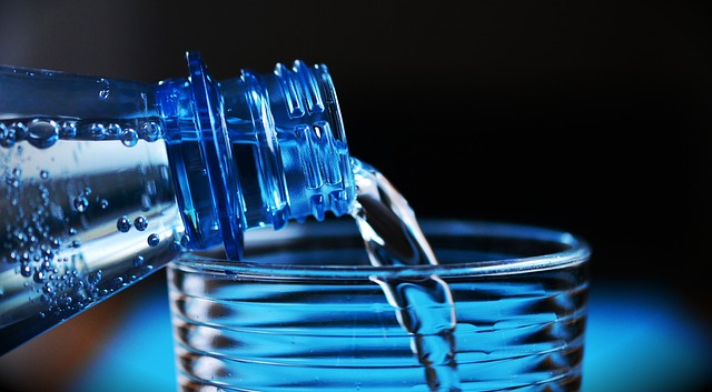 Er wordt water uit de fles in het glas gegoten. Alleen de hals van de fles en de bovenkant van het glas zijn zichtbaar. De achtergrond is donker en blauw.