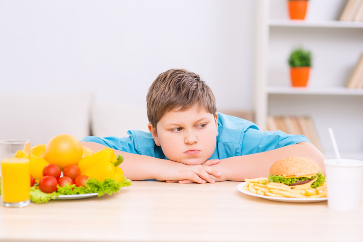 Een dik kind tussen twee borden met gezond en ongezond eten.