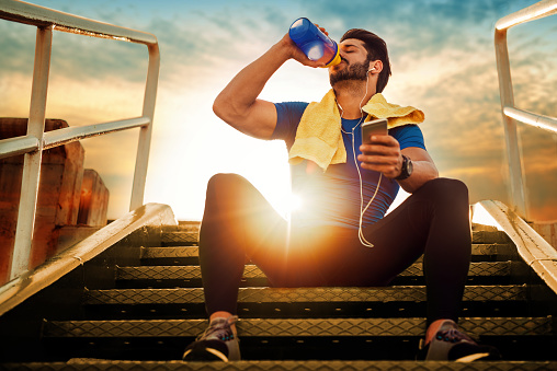 Een atleet die op de trap zit. Zonsondergang. Een voedingssupplement uit een potje drinken.