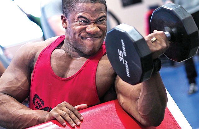 Man in rode tanktop. Hij doet biceps met een eenarmige barbell.