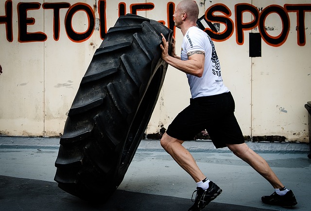 Een man in een wit T-shirt en een zwarte korte broek is aan het trainen. Hij gooit een grote tractorband voor zich om.