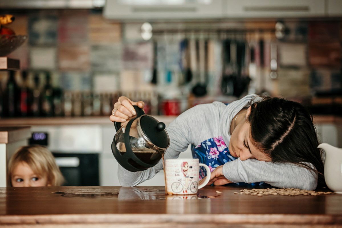 Een vermoeide vrouw ligt op het aanrecht, koffie te morsen naast een mok, een kind staat naast haar.