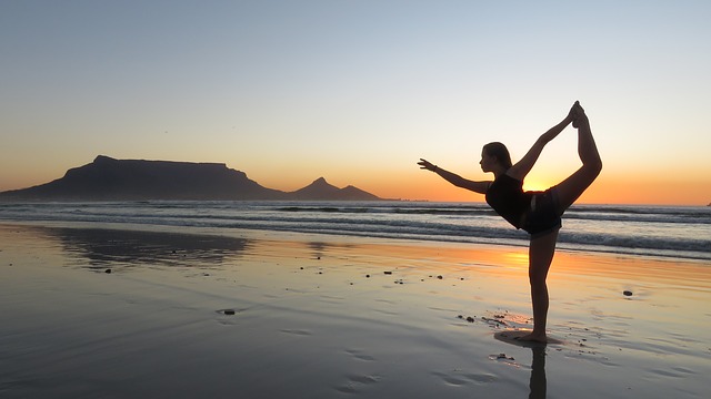 Een vrouw doet aan yoga op het strand. De zon is onder, het is nog licht. Ze staat op één been. Het is eb. Het zand ligt onder een dun laagje water.