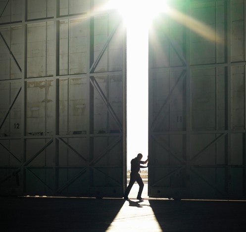 Een man loopt uit de donkere hal het felle licht in, de zon schijnt, hij opent een deur in de hal